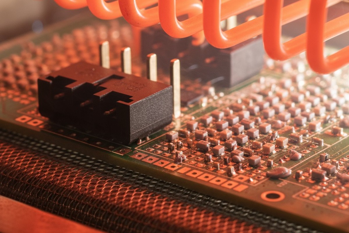 A macro view of a printed circuit board with various electronic components moving on a conveyor belt inside a reflow oven with glowing orange heating elements.