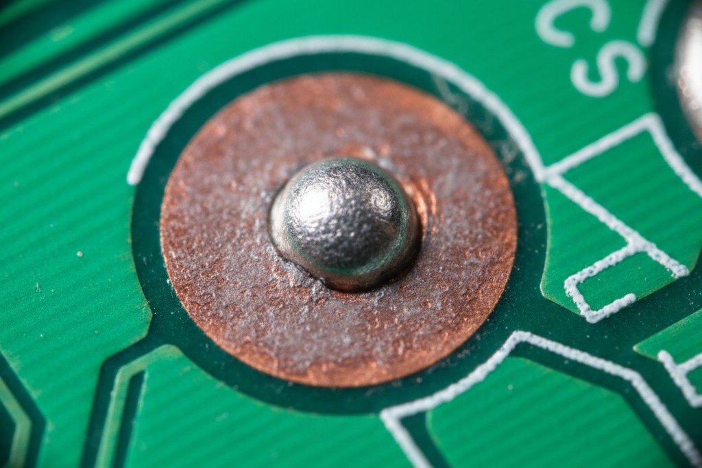 A macro photo of a PCB showing a failed solder joint where the solder has balled up on a dull, oxidized copper pad.