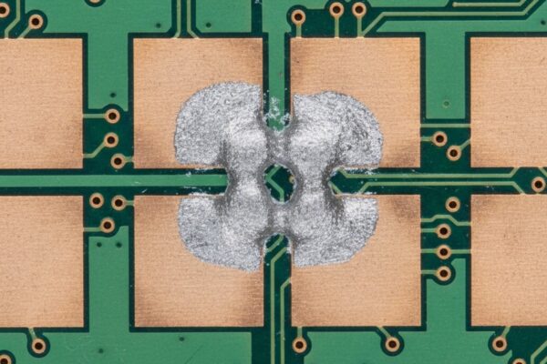 An extreme close-up, top-down view of a green printed circuit board shows a gray solder paste bridge that has formed between two adjacent copper pads, illustrating a cold slump defect.