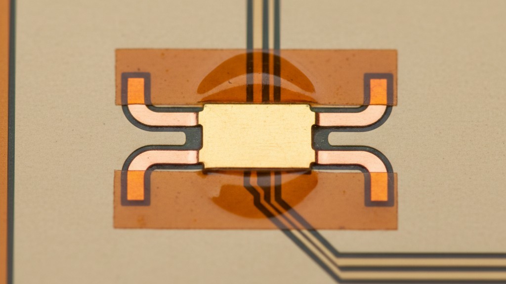 A close-up of a flexible circuit shows a rectangular copper pad with smaller copper extensions, or spurs, that reach under the amber-colored coverlay to secure it.