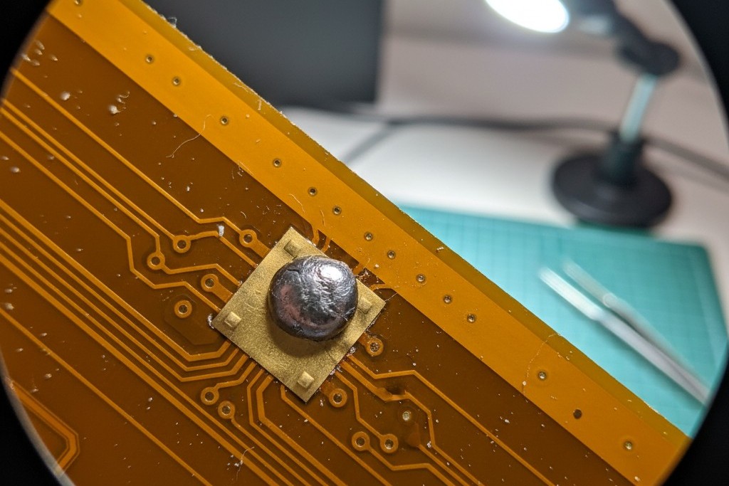 A macro photograph of a gold-plated pad on a flexible circuit board. A sphere of solidified solder sits on the pad instead of flowing across it, indicating a soldering defect.