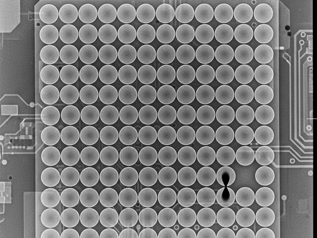 A monochrome X-ray image showing the grid of solder balls beneath a Ball Grid Array (BGA) chip, revealing a subtle 'head-in-pillow' defect on one of the connections.