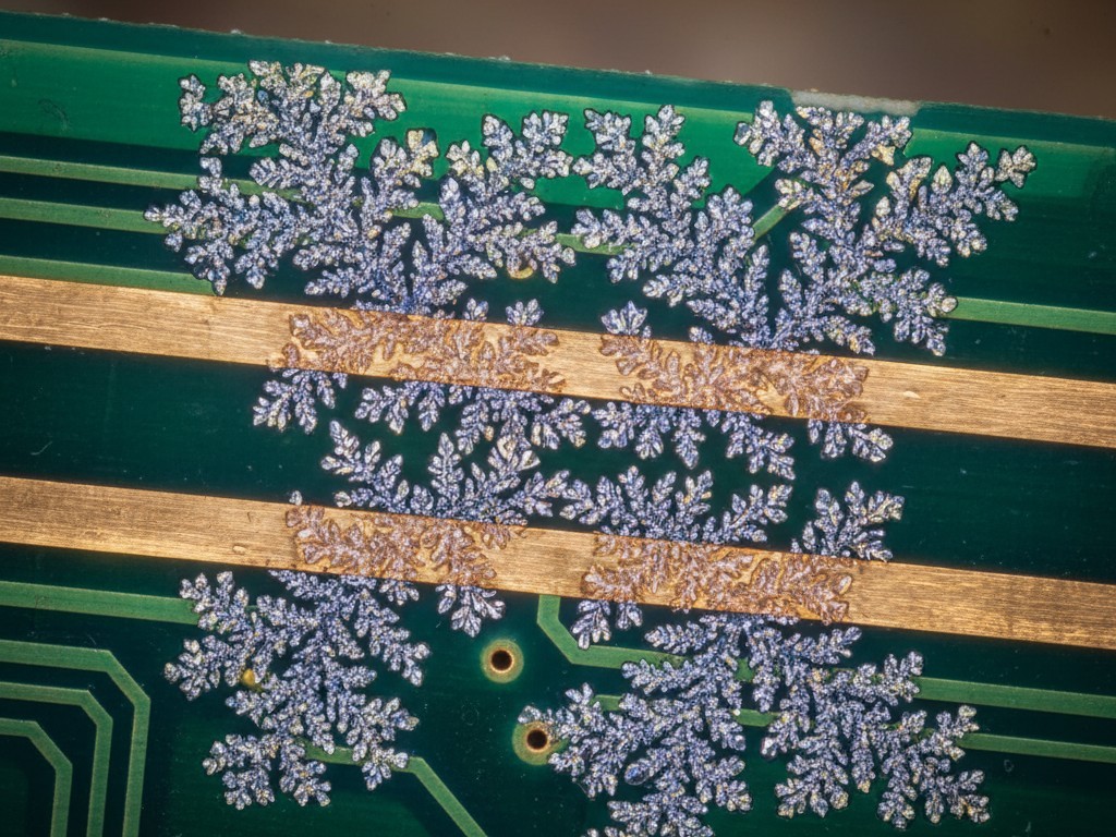 Microscopic dendritic growth on PCB A magnified view of a printed circuit board showing fern-like metallic crystals growing between two copper traces.