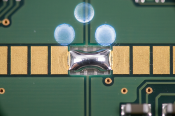 Solder Mask Expansion: The Invisible Yield Killer A magnified circular view of a green PCB reveals a silver solder bridge connecting two adjacent gold pads, illuminated by three bright circular reflections.